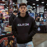See the 'Glitch' men's black hoodie by Raverge in action, worn by a model. The design features a unique glitched text effect, making it a standout piece for fans of digital aesthetics and gaming culture. With its distorted typography, this hoodie brings a modern and edgy vibe to streetwear, perfect for those who love to make a statement with their style.