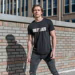 Model wearing a black "UNLEASHED" t-shirt with bold typography and a red slash, posing against a brick wall.