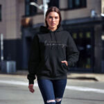 Model wearing Raverge women's black hoodie with 'Embrace Harmony' text in stylish script.