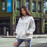 Model wearing Raverge women's white hoodie with 'Embrace Harmony' text in stylish script.