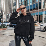 Model wearing Raverge women's black hoodie with 'Glam Girl' text in an elegant design.