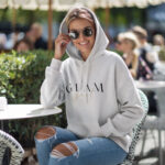 Model wearing Raverge women's white hoodie with 'Glam Girl' text in an elegant design.