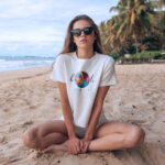 Model wearing Raverge Women's Beach Chic T-Shirt with a beach ball graphic, sitting on sandy beach