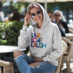 Model wearing Raverge women's white hoodie with "Live Love Laugh" colorful typography design.