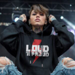 Model wearing Raverge women's black hoodie with "Loud 'N Proud" design and red lightning bolt for rock music fans.
