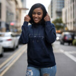 Model wearing Raverge women's navy hoodie with 'Embrace Harmony' text in stylish script.