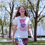 Model wearing a "Love is All You Need" women's t-shirt with colorful, retro-style text, set against a relaxed backdrop.