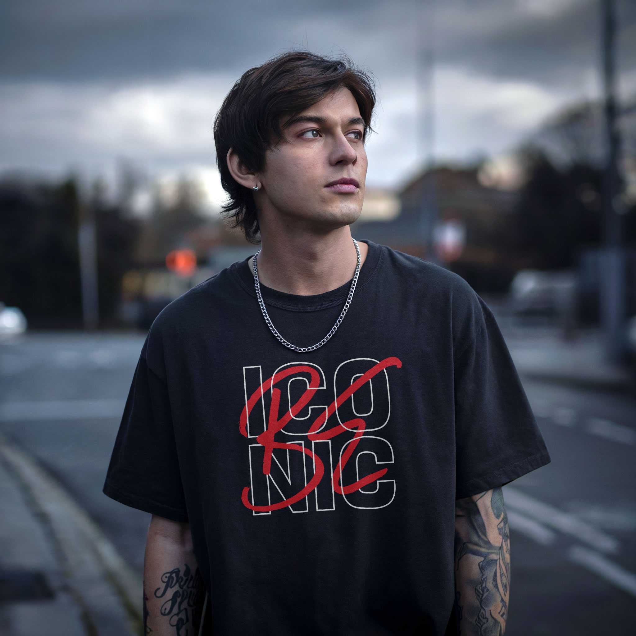 Stylish young man wearing a black oversized streetwear t-shirt with "BE ICONIC" typography, standing confidently in an urban setting.