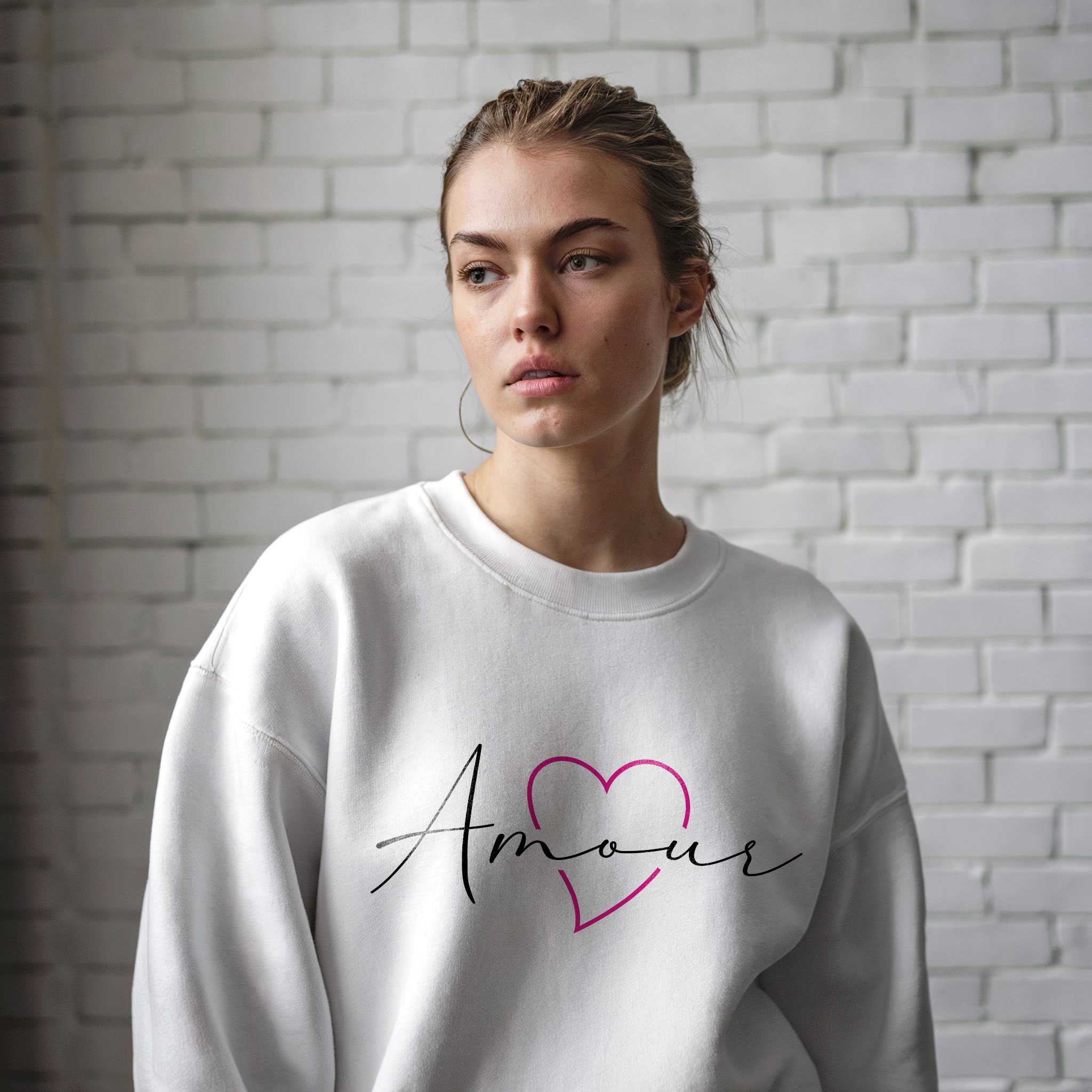 Trendy young woman wearing a white oversized sweatshirt with “Amour” typography and a heart design, posing against a brick wall.